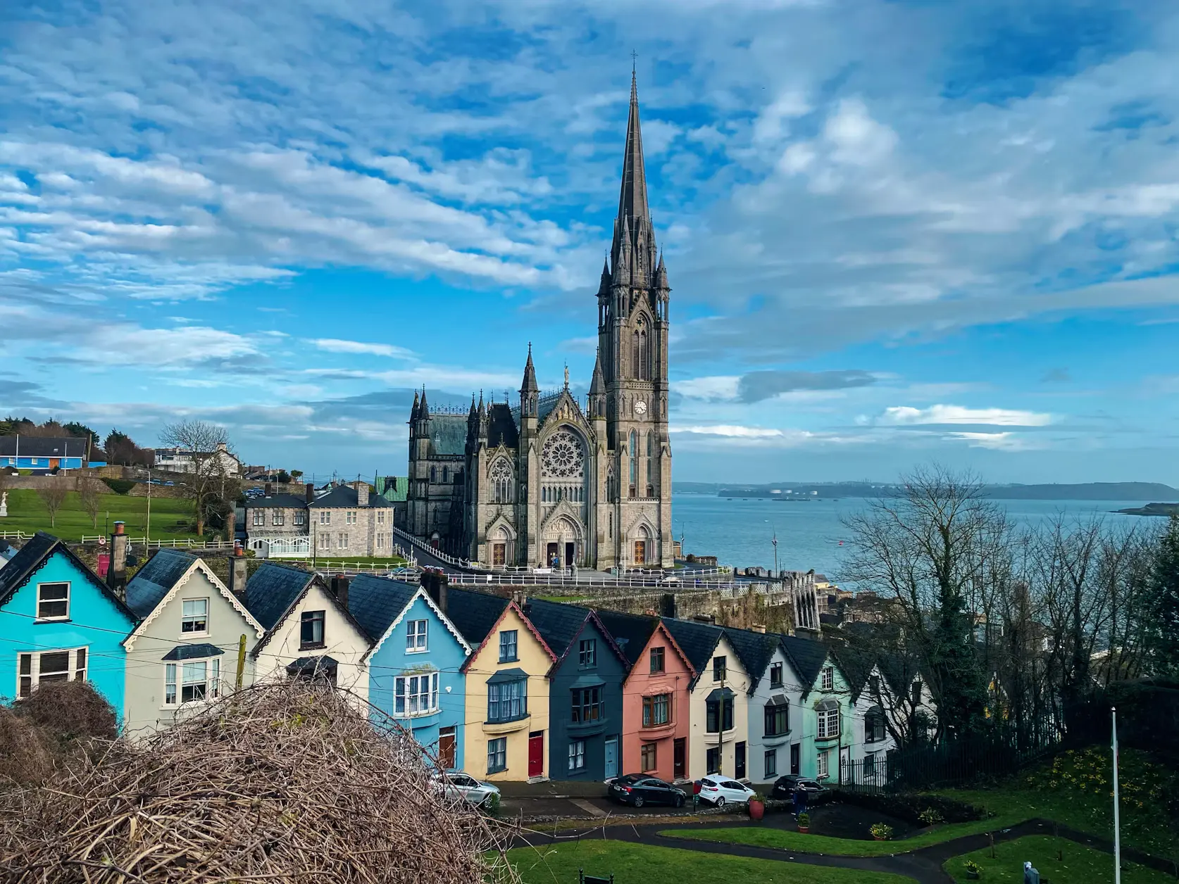 Colorful row houses on a hillside overlooking a harbor town, with a tall Gothic cathedral spire rising above, Cobh, Ireland.