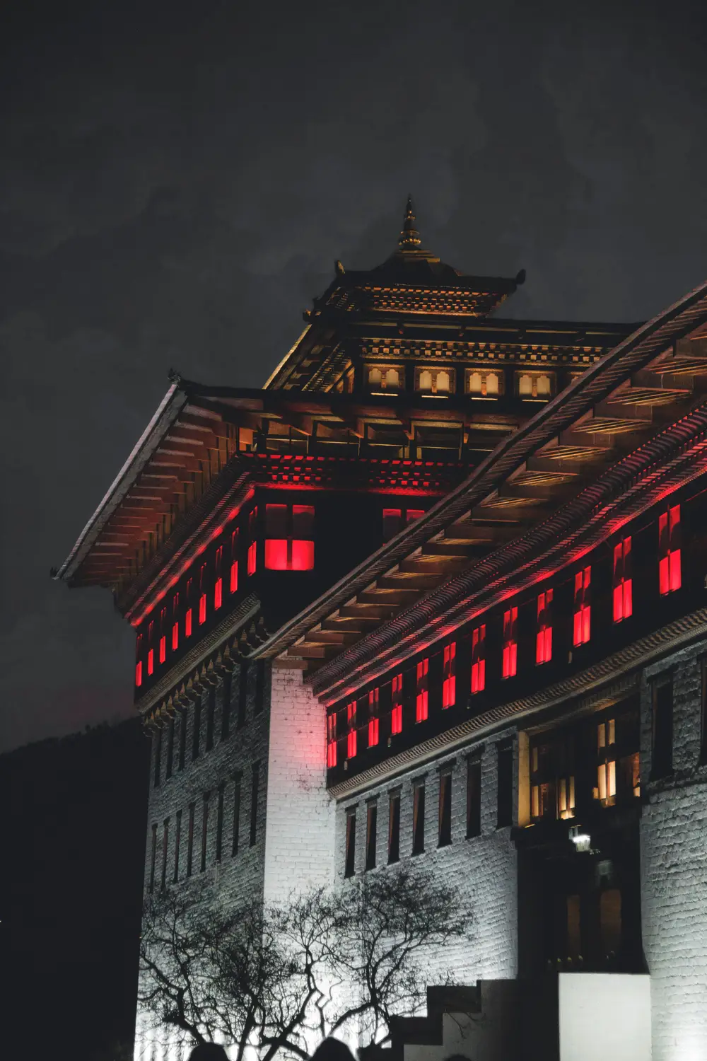 Traditional Bhutanese dzong illuminated at night, showing ornate red-lit windows and tiered architecture against a dark sky in Bhutan.