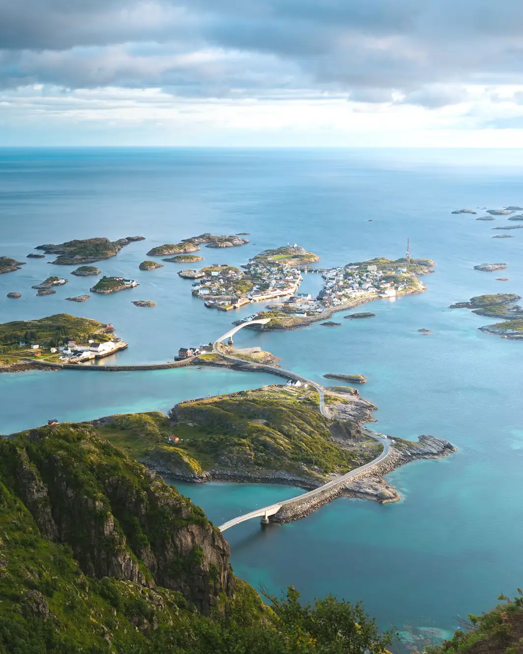 Aerial view of Henningsvær village spread across small islands connected by bridges, surrounded by the blue waters of the Norwegian Sea.