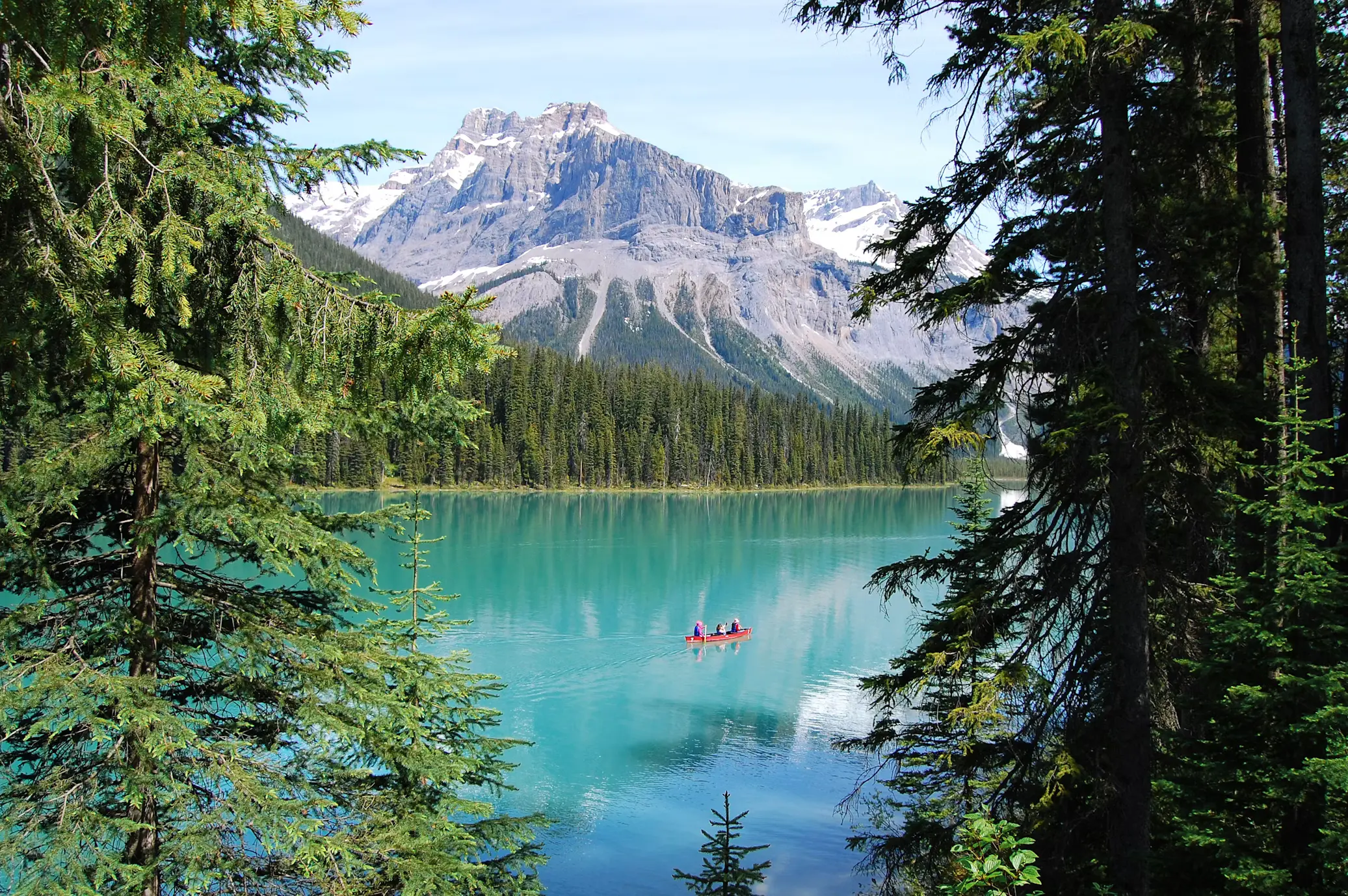 A turquoise glacial lake framed by pine trees, with small red canoes on the water and snow-capped mountains in the distance.