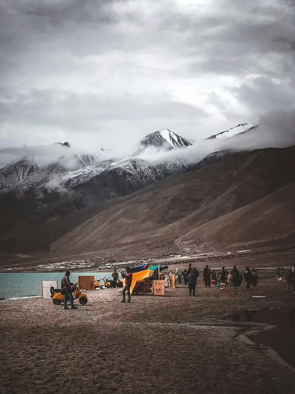 Tourists at Pangong Tso lake shore with snow-capped Himalayan mountains and dramatic storm clouds, Ladakh, India
