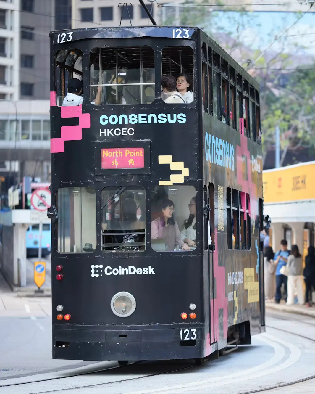 Double-decker tram with colorful advertising wraps traveling through a busy city street in Hong Kong.