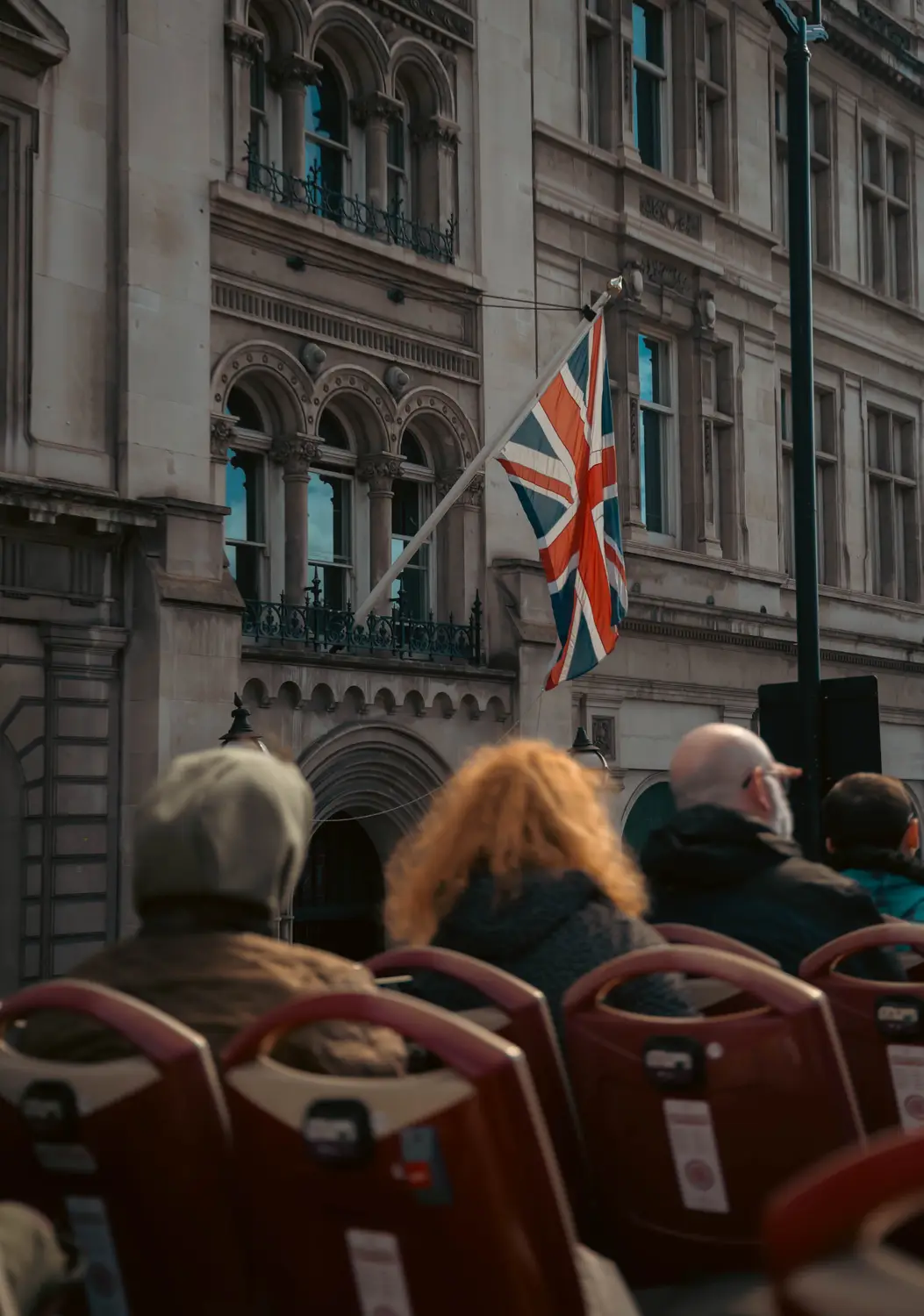 View from the top deck of an open-top tour bus, with passengers ahead and a Union Jack flying from a stone building