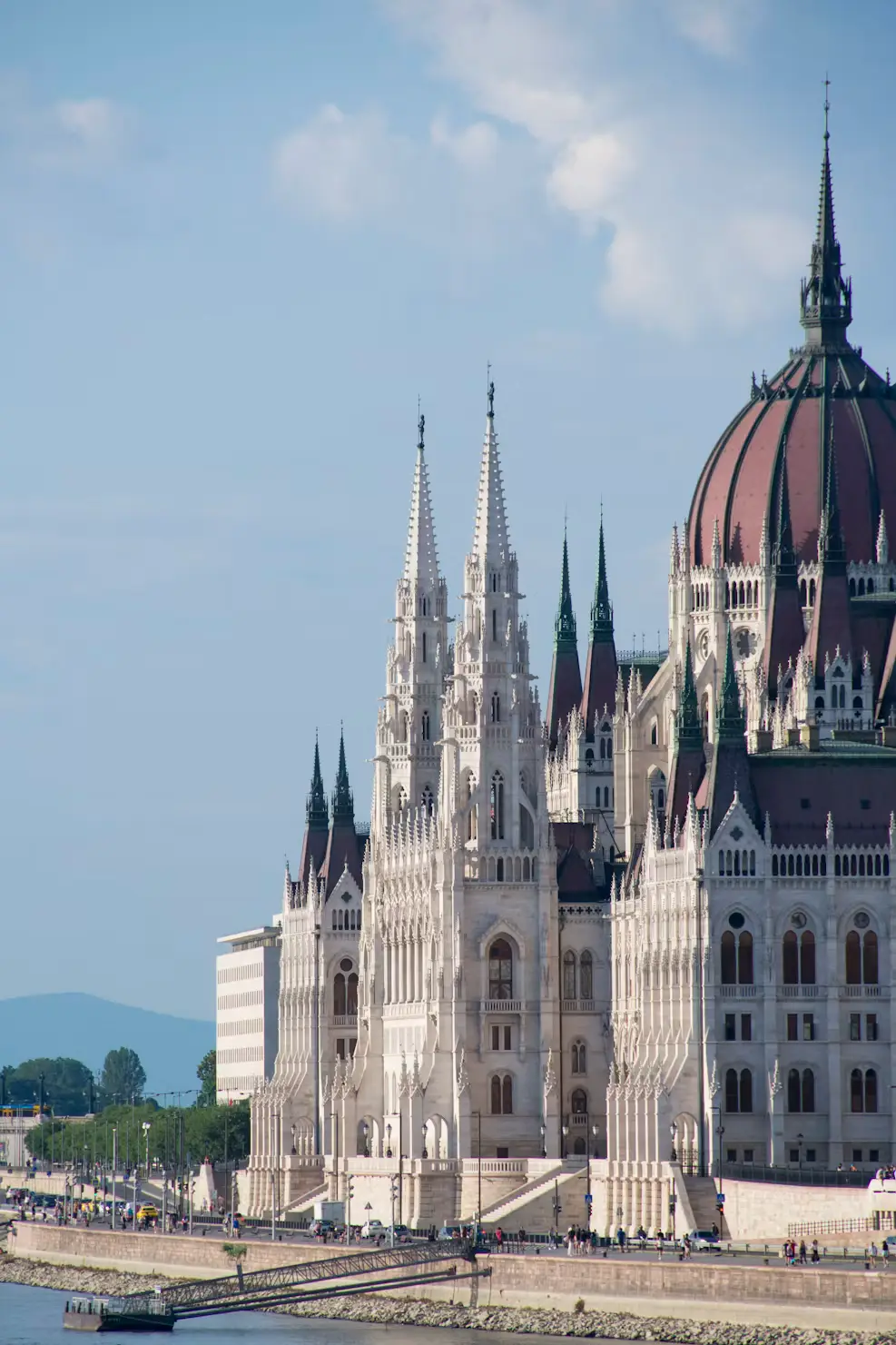 Ornate white parliament building with tall Gothic spires and a large dome along a riverside promenade.