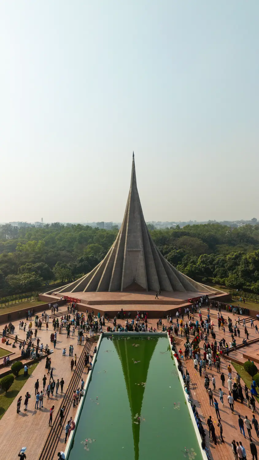 Aerial view of National Martyrs' Memorial (Jatiyo Smriti Soudho) in Savar with crowds of visitors, reflective green pool, and surrounding green trees, Dhaka, Bangladesh