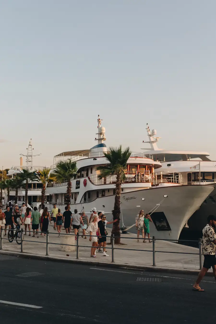 Large white yacht docked along a waterfront promenade with people walking nearby at dusk.