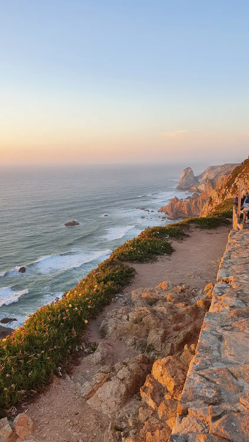 Rocky coastal cliffs overlooking the ocean at sunset, with waves below and a stone lookout path along the cliff edge.