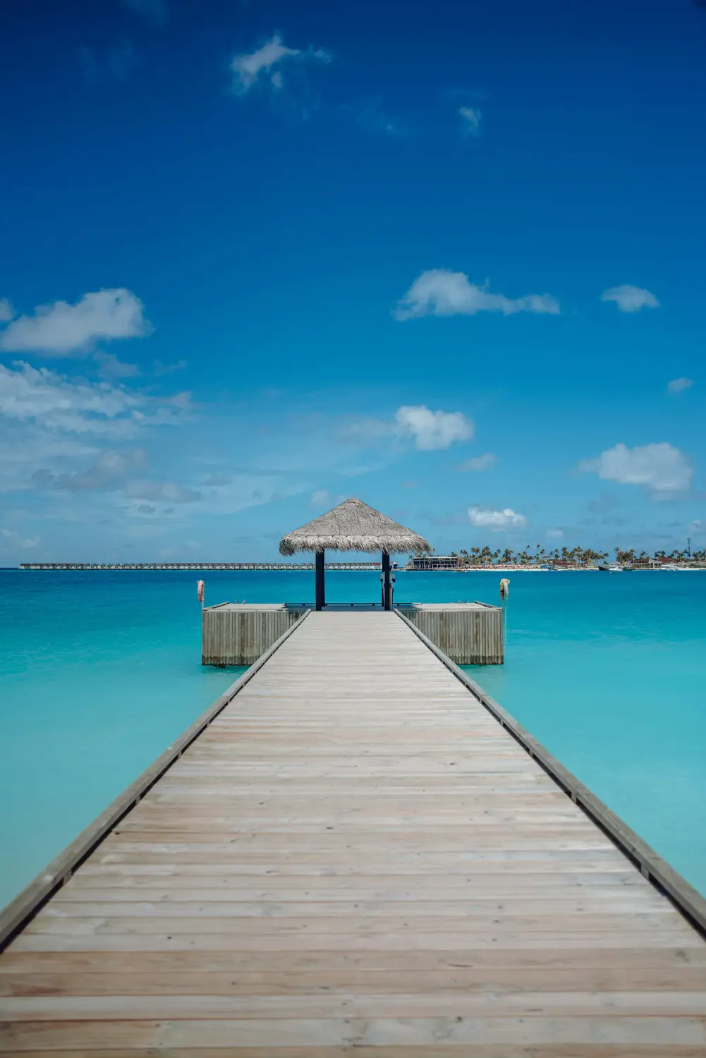 Straight wooden boardwalk jetty extending over crystal clear turquoise lagoon to overwater bungalow pavilion under blue sky in Maldives