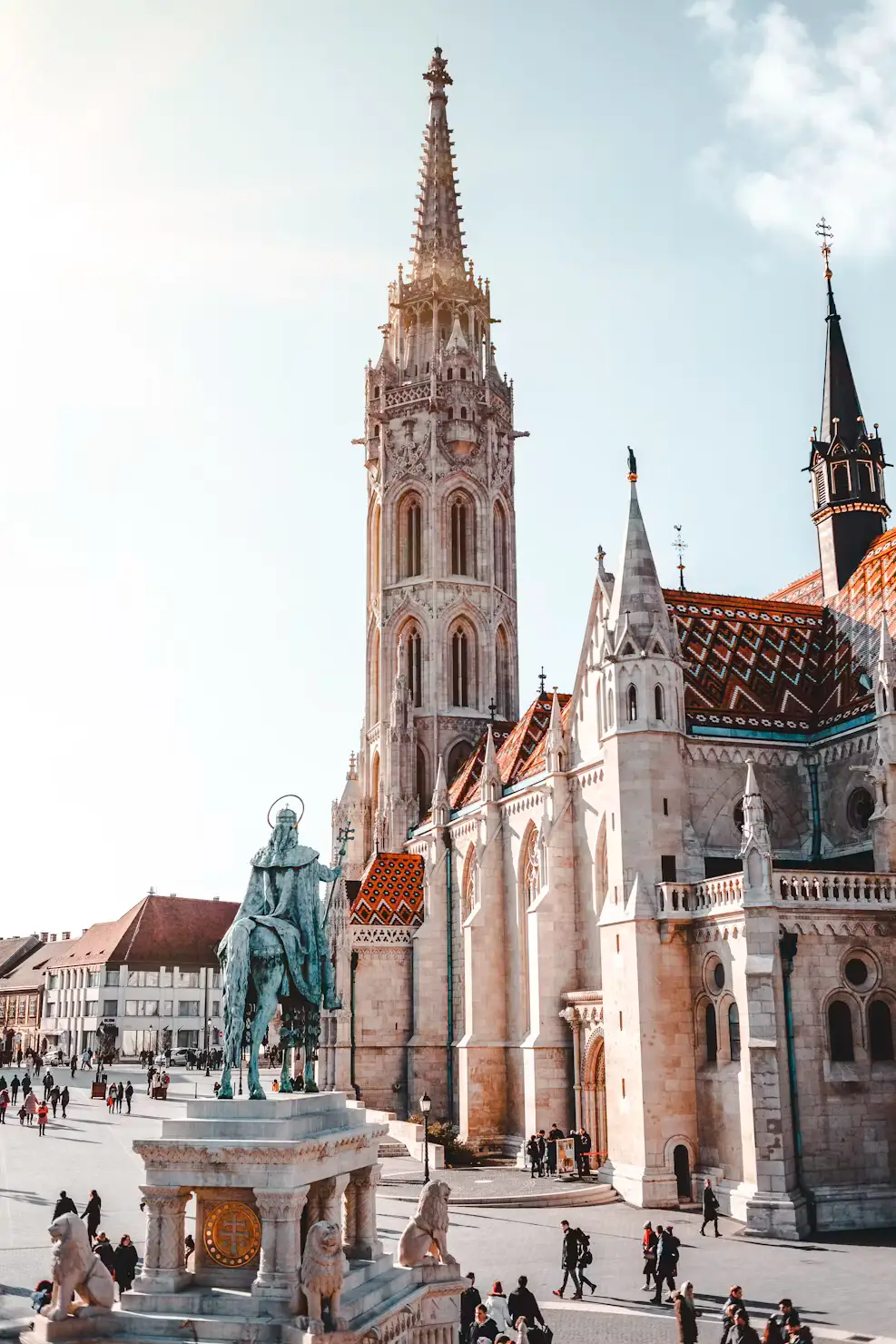 Gothic church with a tall ornate spire and patterned roof beside an equestrian statue in a busy city square.