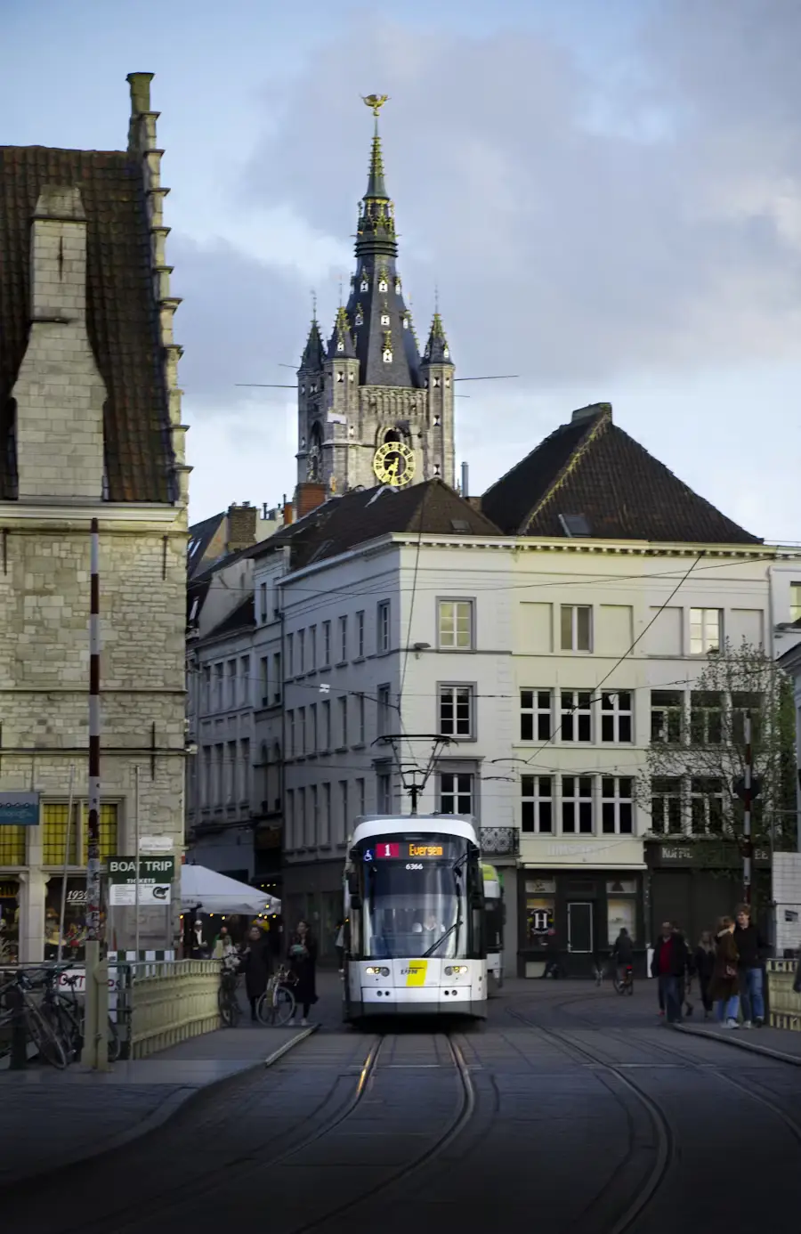 Tram passing through a historic Belgian street with a Gothic belfry tower rising in the background at dusk