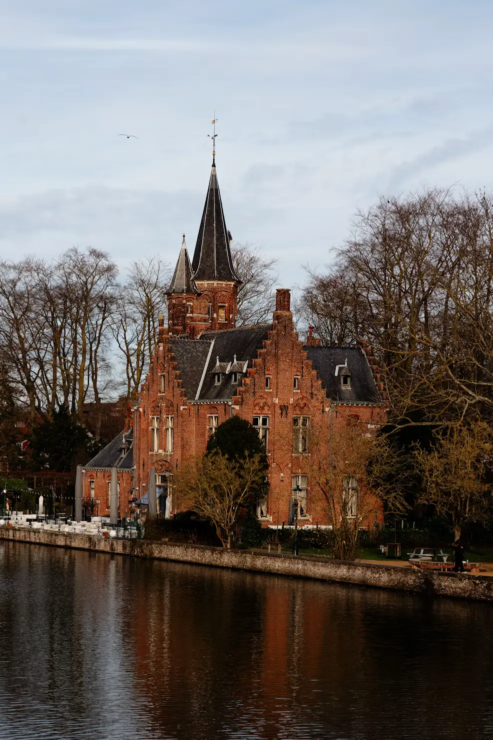 The red-brick Lock House (Minnewater) with its steep spire reflected in the water, surrounded by bare trees in Bruges.