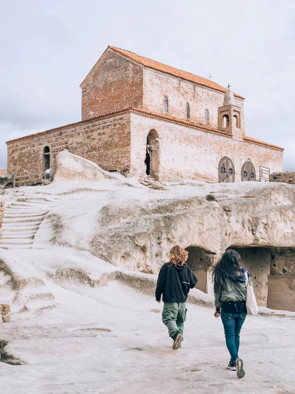 A weathered stone-and-brick church atop white rock, with two people walking toward it.
