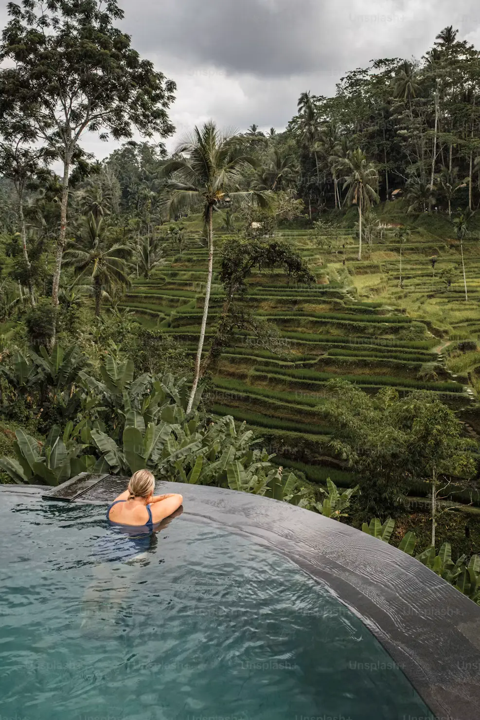 Person relaxing in an infinity pool overlooking terraced rice fields and tropical jungle in Bali, Indonesia.