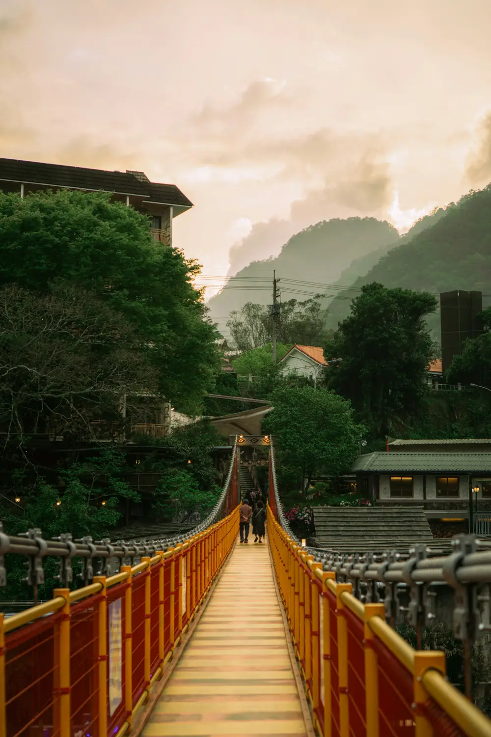 Red and yellow pedestrian suspension bridge leading toward misty green mountains at sunset in Wulai, Taiwan