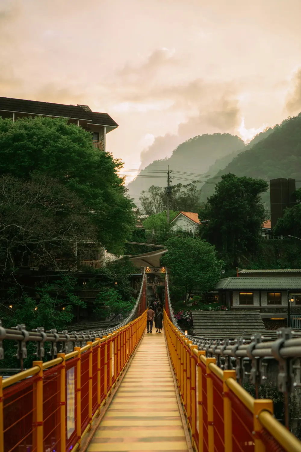 Red and yellow pedestrian suspension bridge leading toward misty green mountains at sunset in Wulai, Taiwan