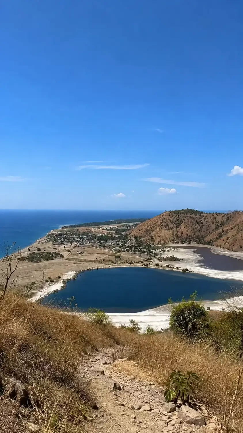 View of Lake Tasitolu in Dili, Timor-Leste from a hilltop hiking trail, showing the serene blue lagoon surrounded by dry hills, coastal wetlands, and the Timor Sea in the background