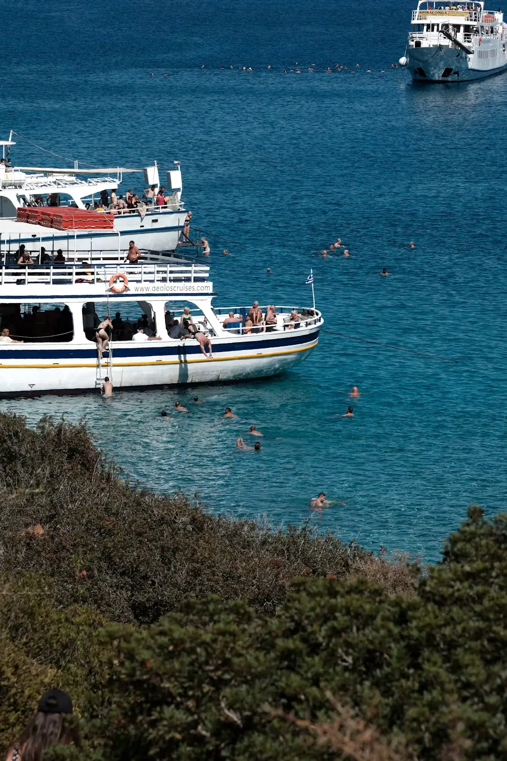 Boats anchored in a clear turquoise cove with people swimming nearby, framed by green coastal shrubs.