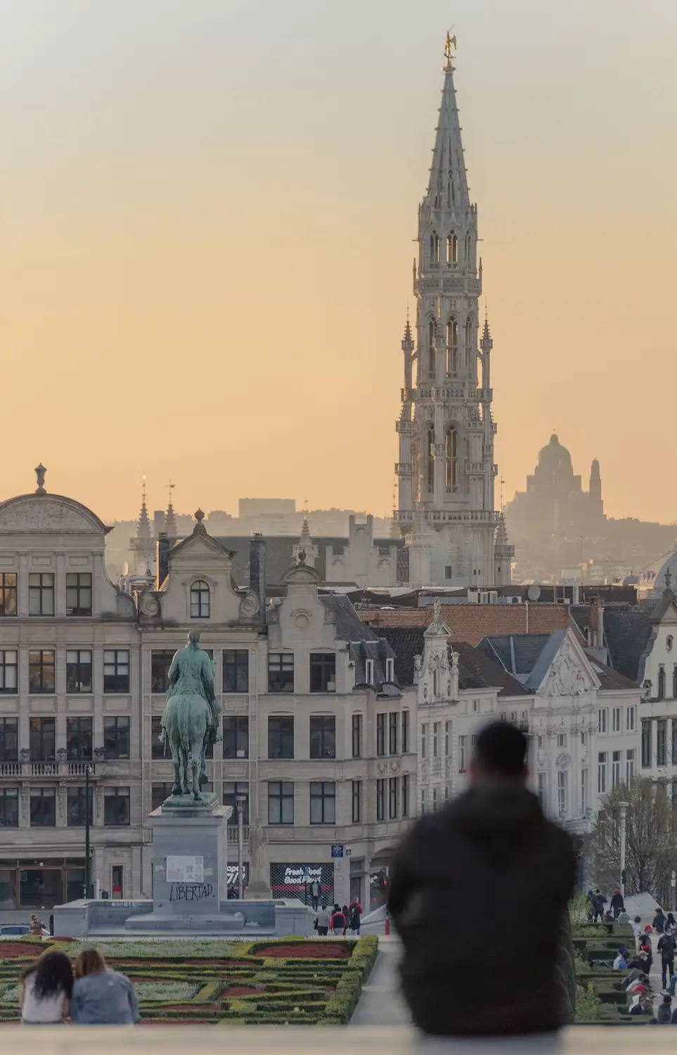 A person overlooking Brussels' skyline at sunset, with the Town Hall spire and a statue rising above the Mont des Arts gardens.