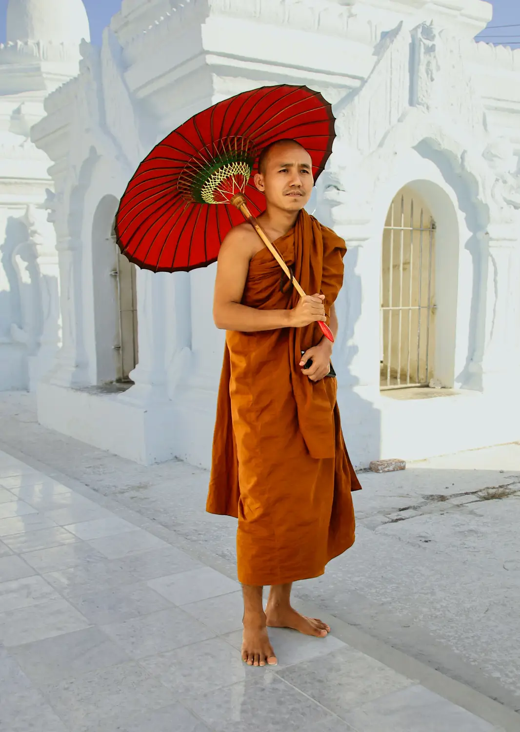 Buddhist monk in saffron robes holding traditional red parasol standing at white Kuthodaw Pagoda, Mandalay Myanmar