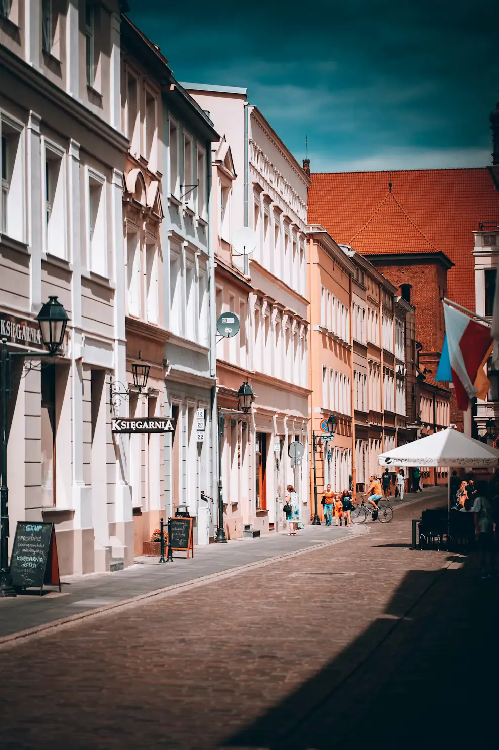 Sunlit street lined with white and orange buildings, with a market stall visible in the distance, Poland.