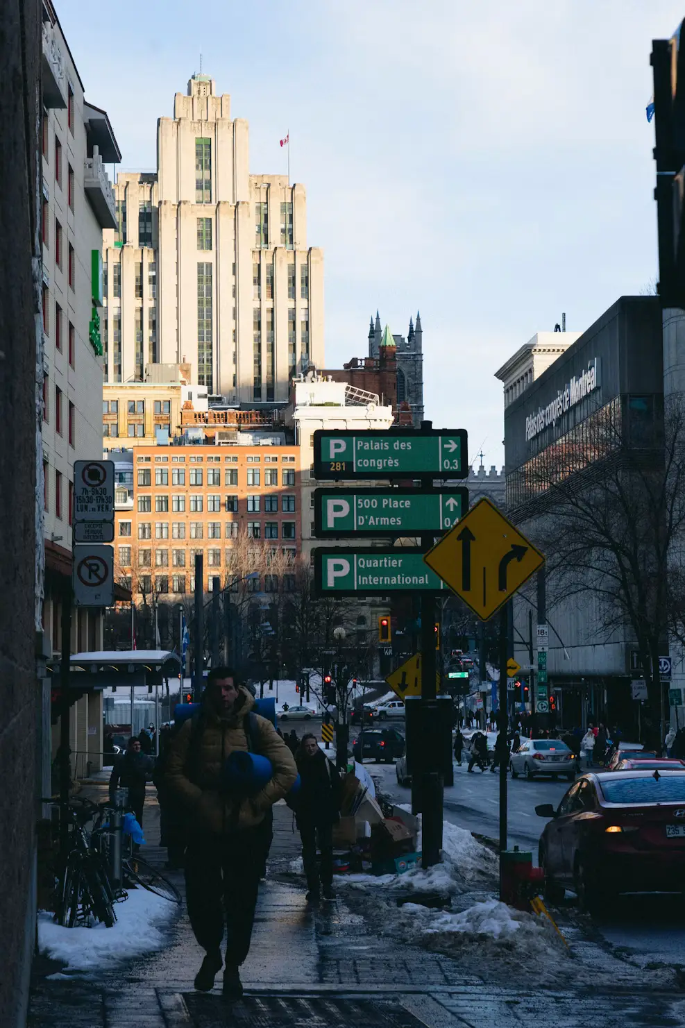 A person walking a dog down a snowy city sidewalk, with a tall historic building and street signs in the background.