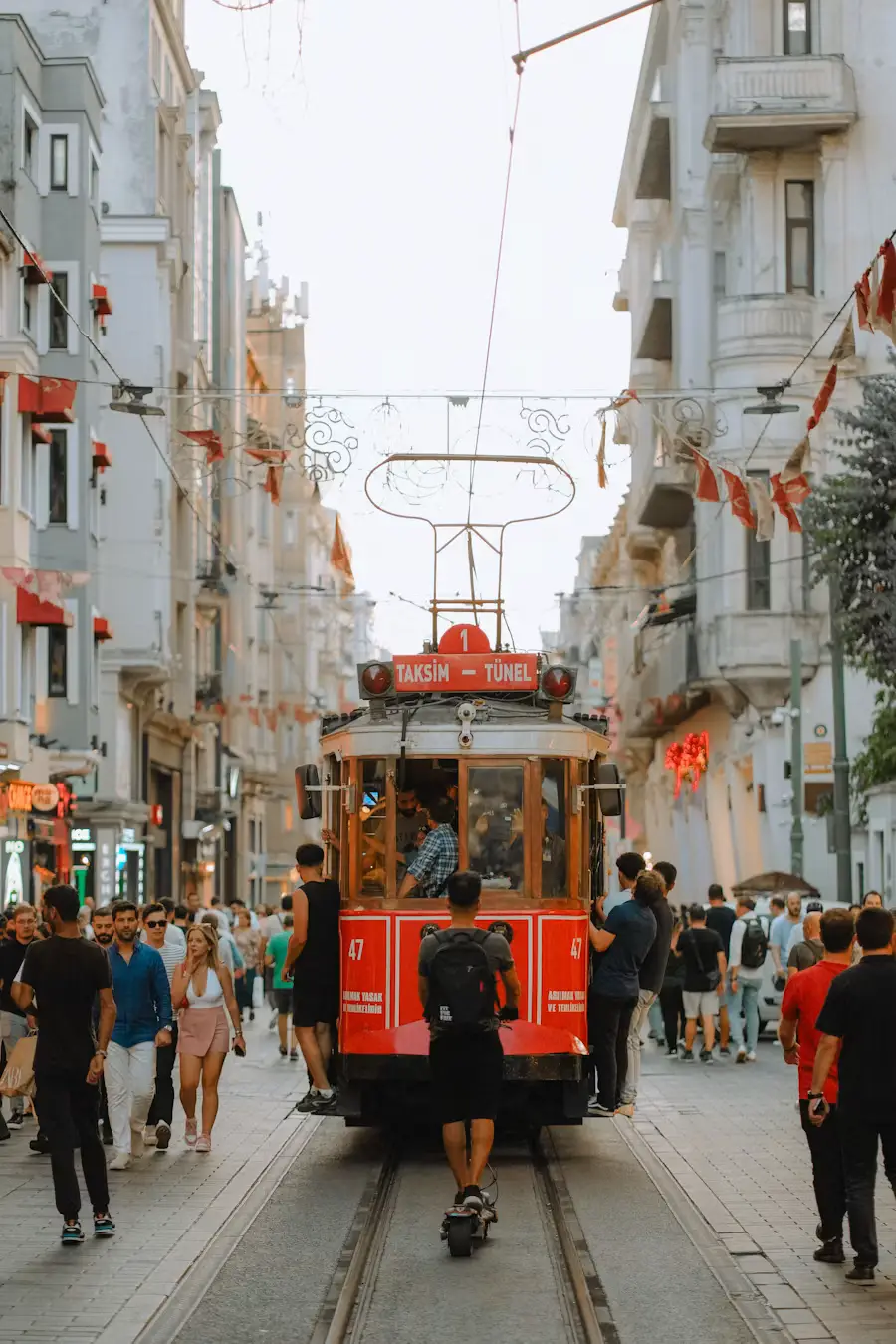 A vintage red tram moving down a busy pedestrian shopping street lined with historic buildings, with crowds of people walking alongside.