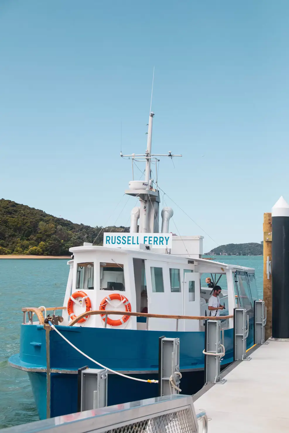 Blue-and-white Russell Ferry boat docked at a pier with calm turquoise water and forested hills behind.
