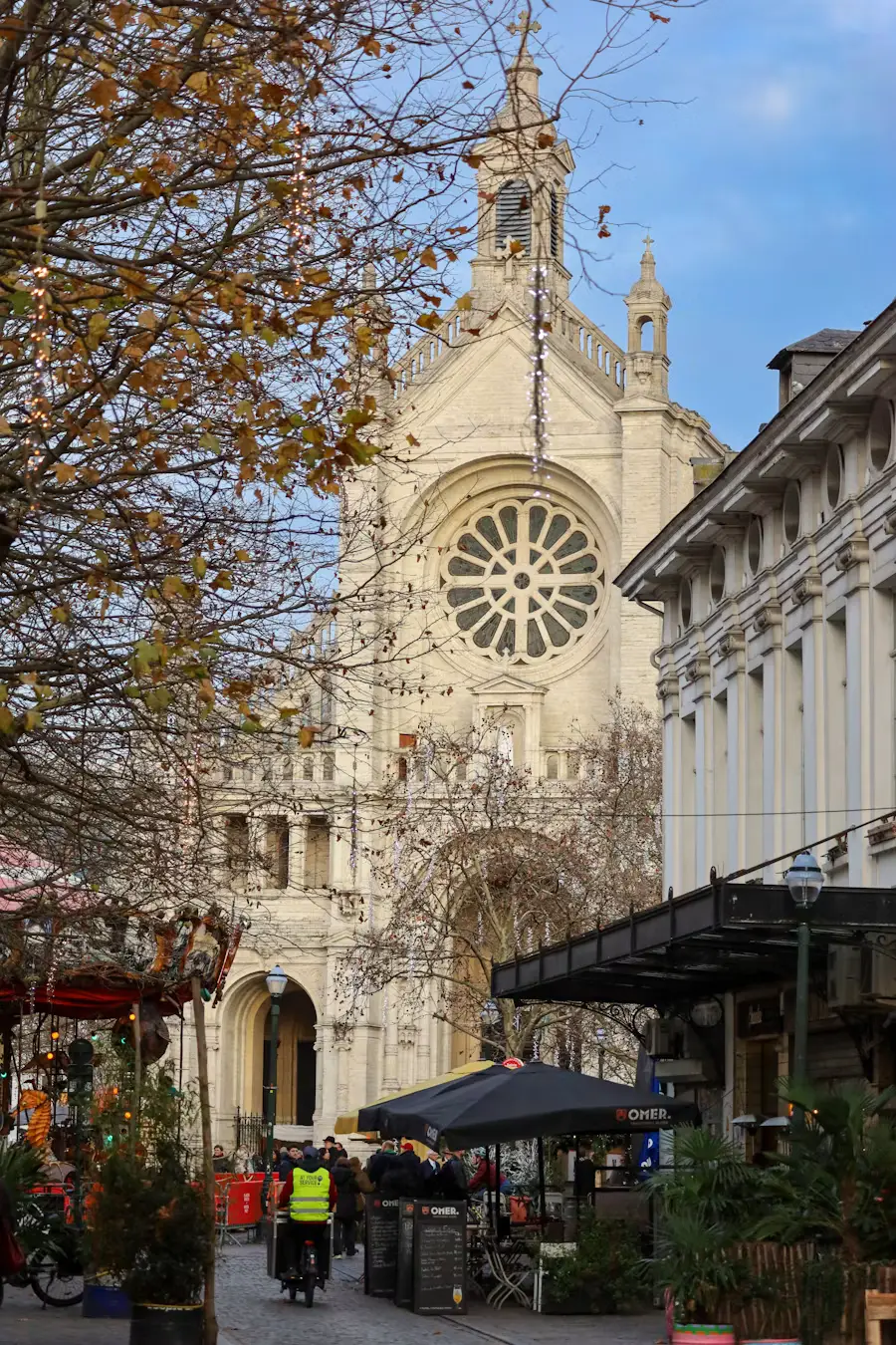 A grand rose-window church facade rising above a lively Brussels street with market stalls and a cyclist passing by.