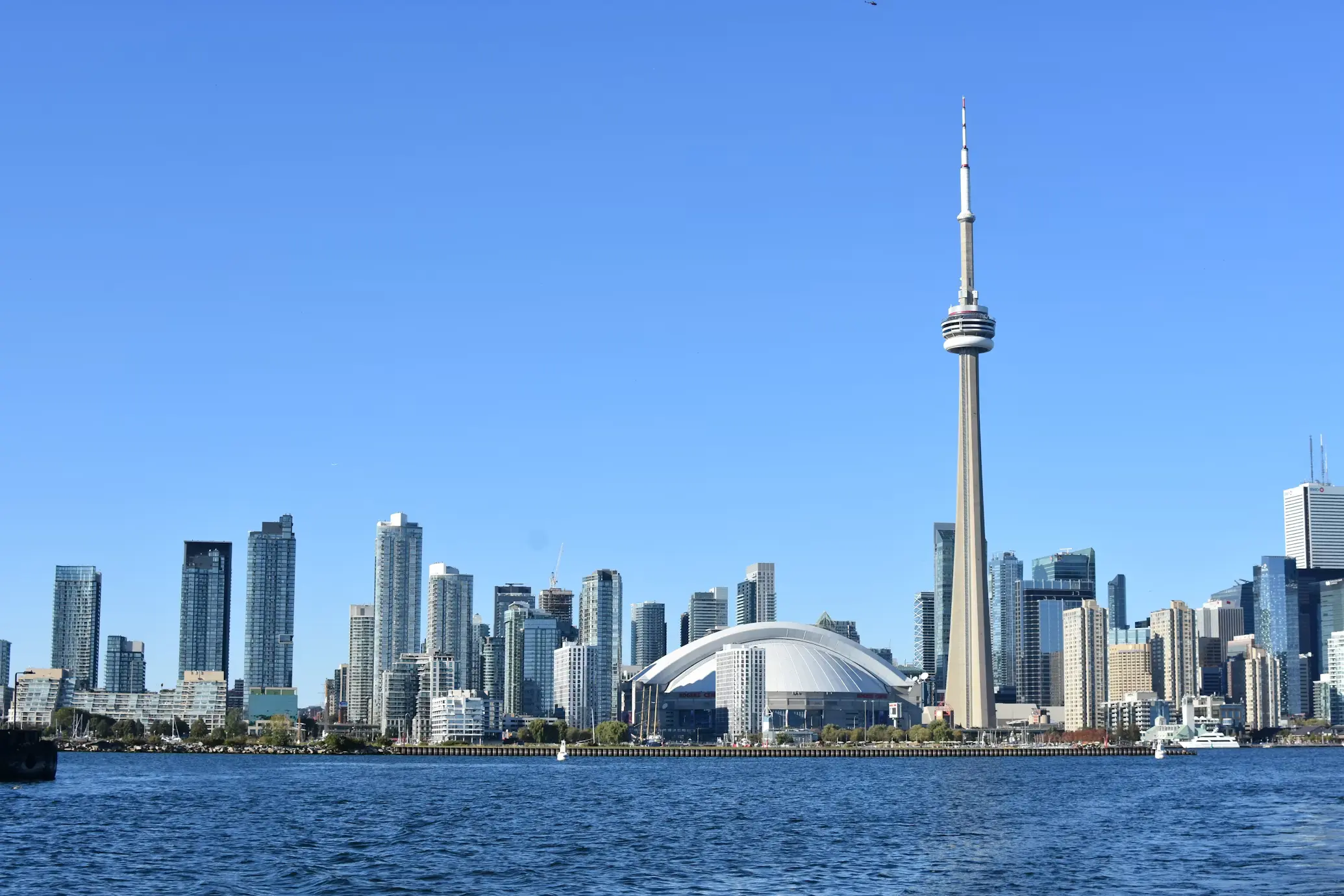 Toronto's skyline and CN Tower seen across the water on a clear day, with sailboats along the waterfront.