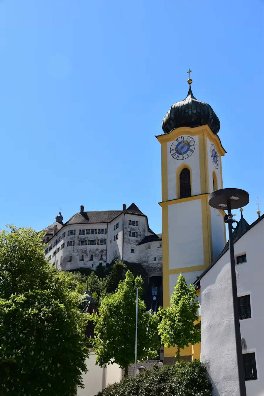 A yellow church tower with a clock and black spire rising above trees, with a hillside building visible behind it under clear blue sky.