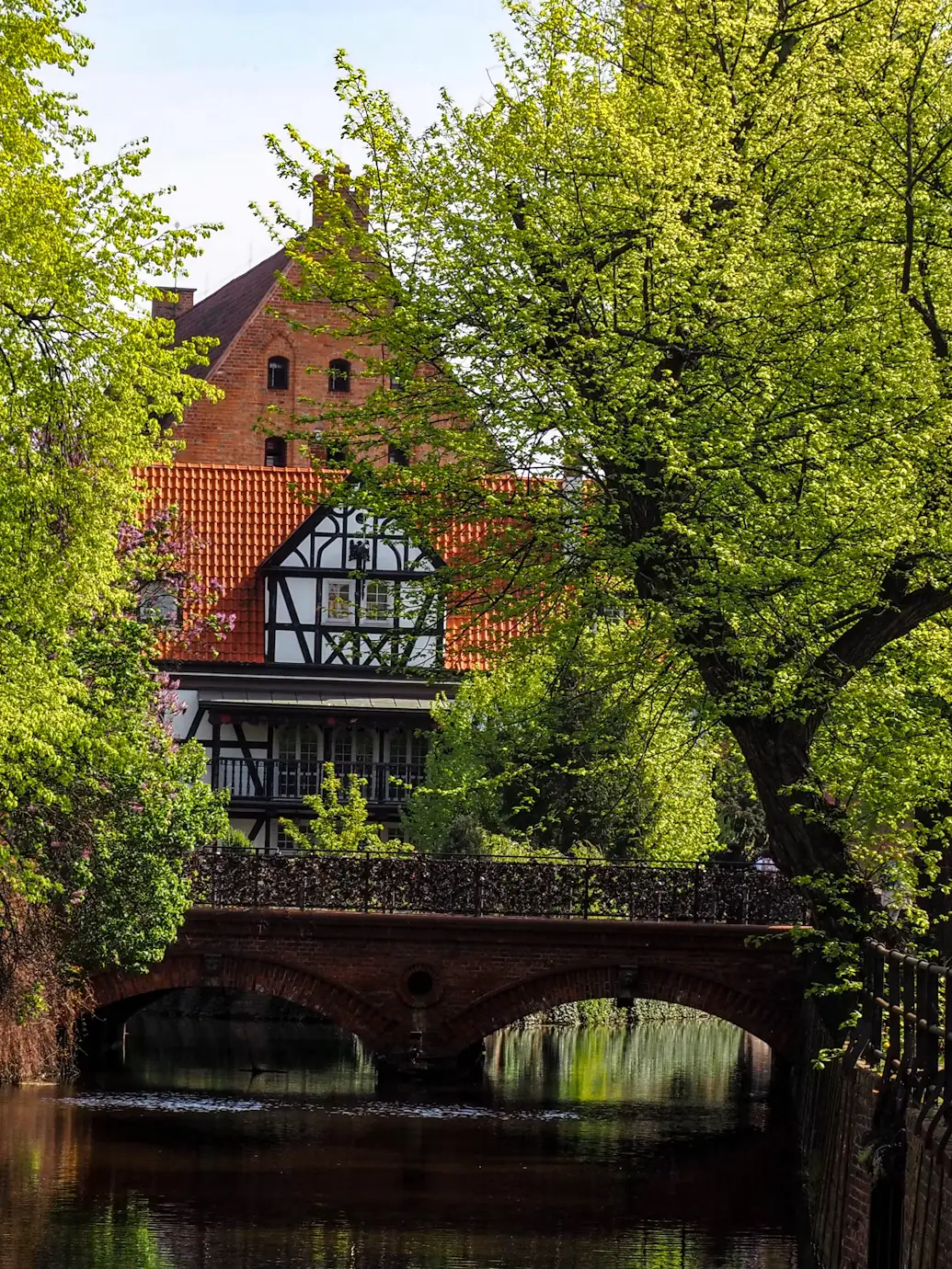 Historic Polish house surrounded by lush green trees, reflected in a calm canal beneath a brick bridge.