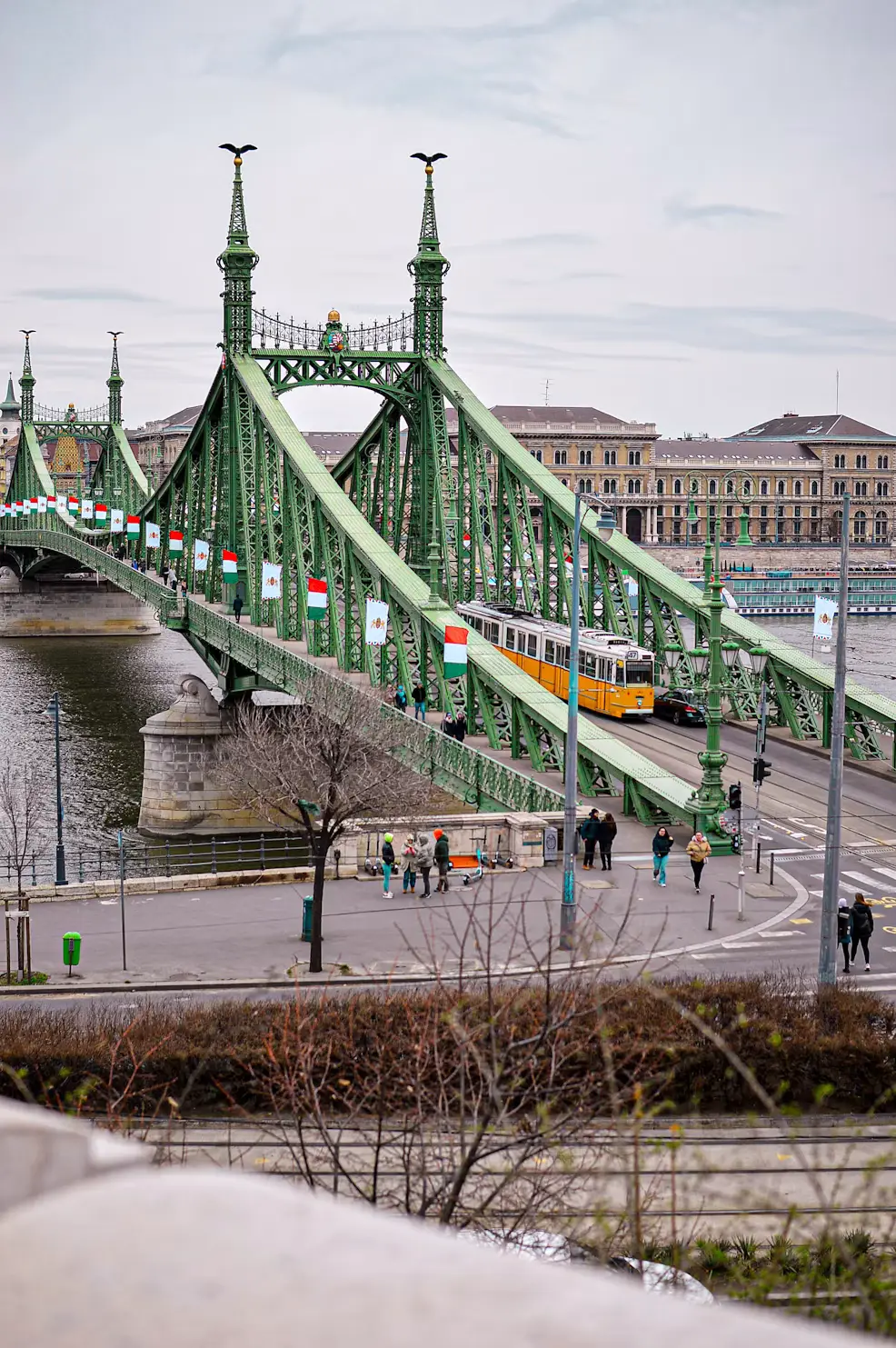 Green steel suspension bridge over a river with a yellow tram crossing and pedestrians along the walkway.