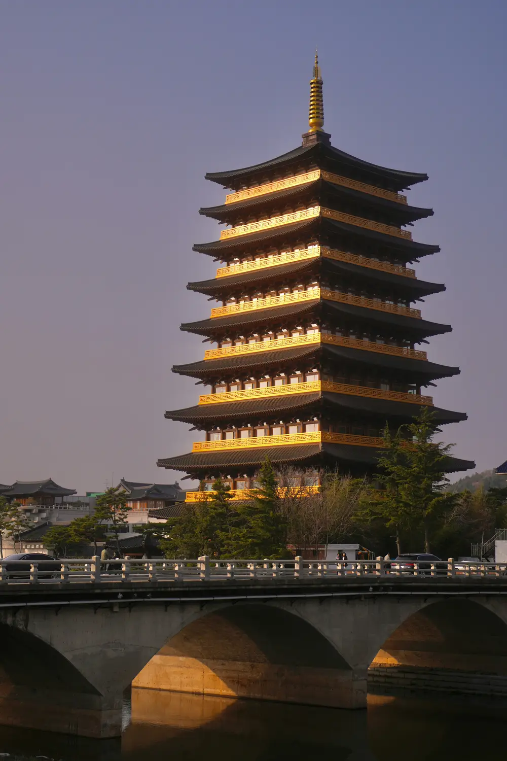 Illuminated multi-story Korean wooden pagoda glowing at dusk beside a stone bridge.
