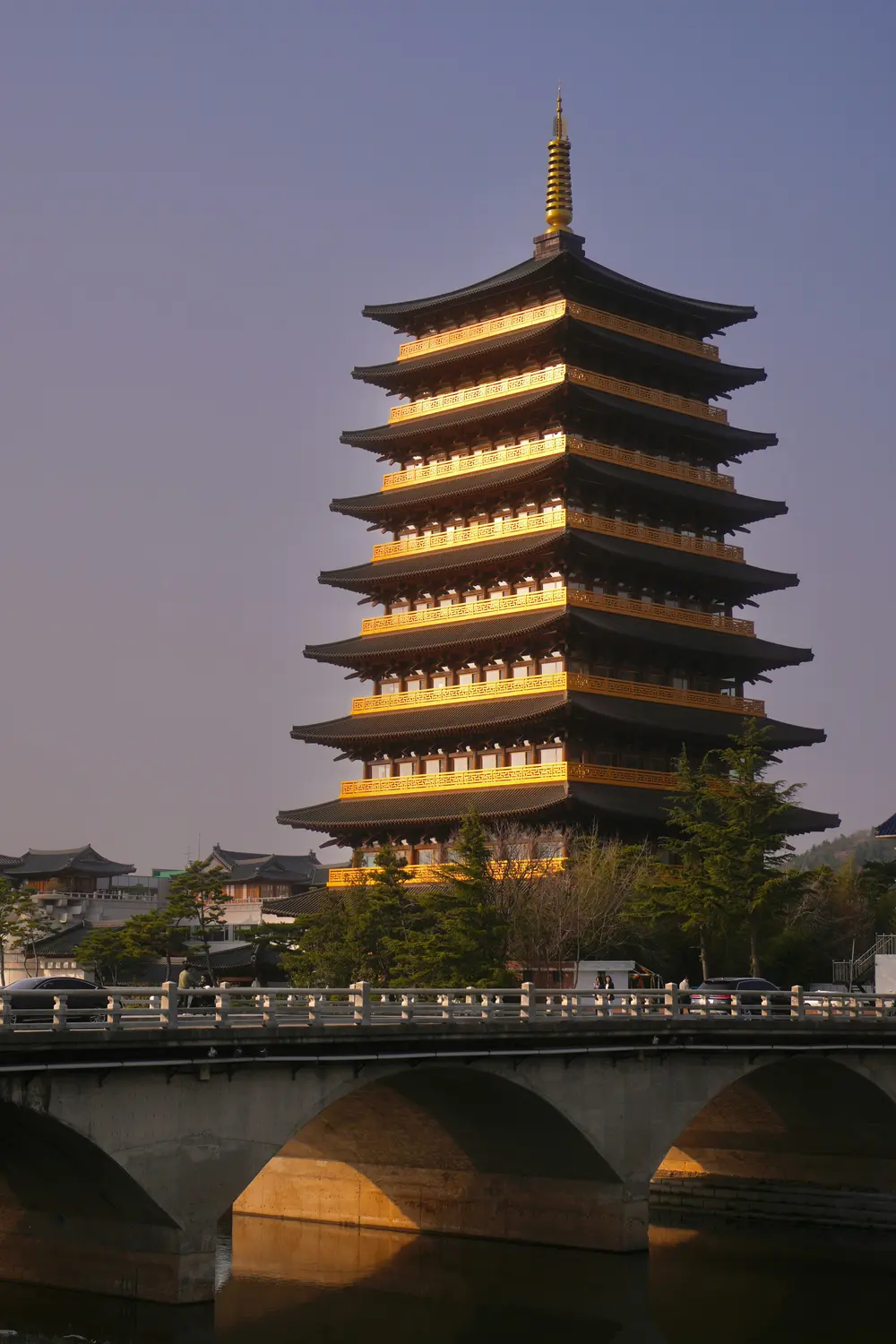 Illuminated multi-story Korean wooden pagoda glowing at dusk beside a stone bridge.