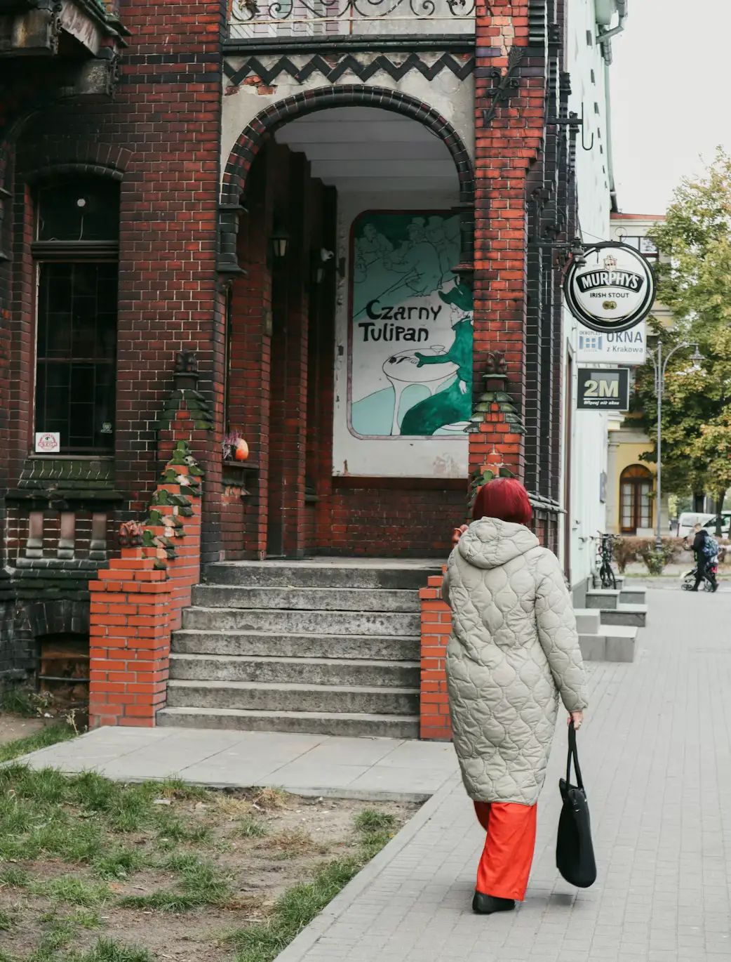 Person in a winter coat walking past a brick building entrance with a striped awning, Poland.