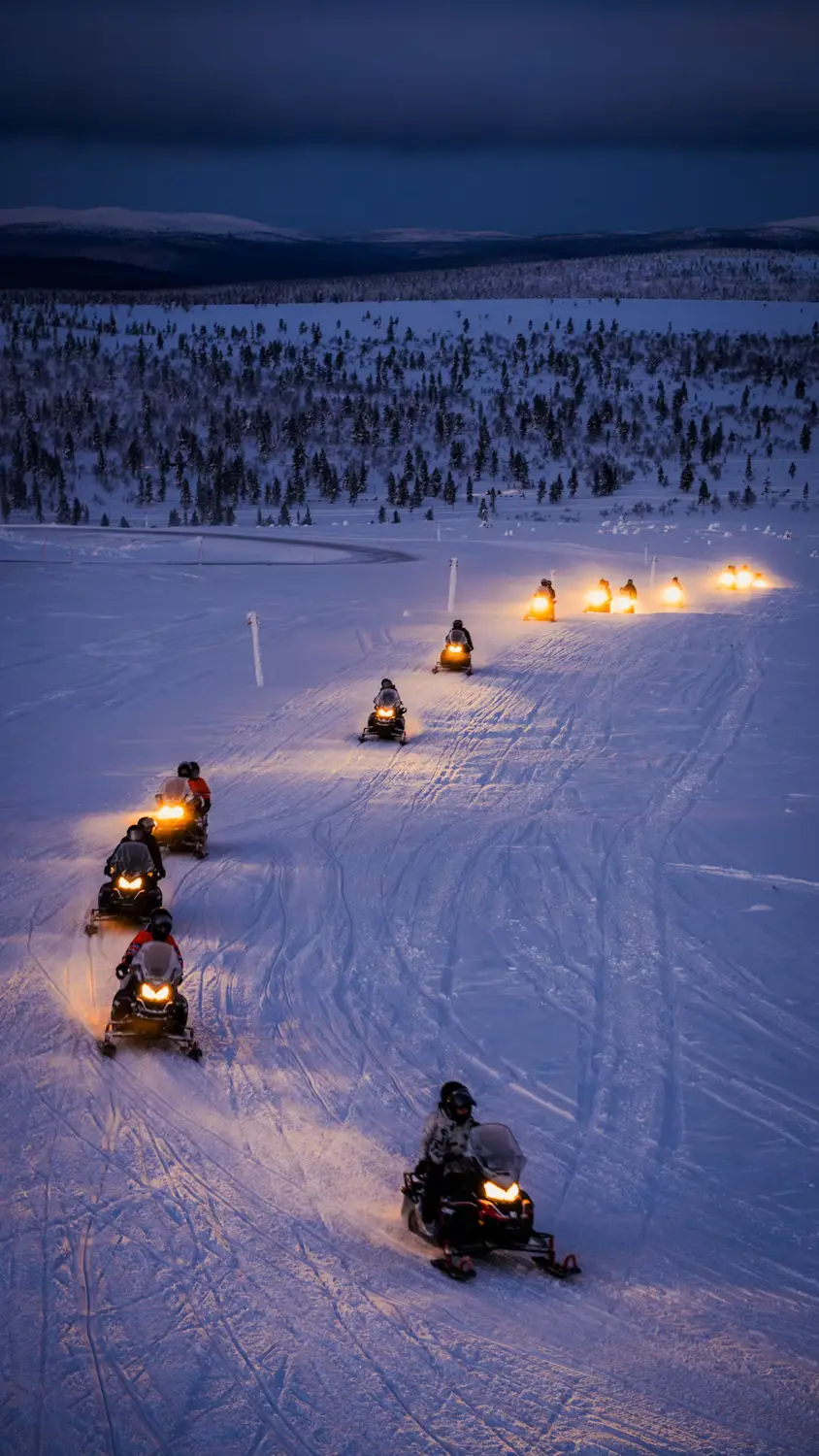 A group of snowmobiles with glowing headlights rides along a snowy trail through a vast winter landscape of forests and distant hills at dusk.