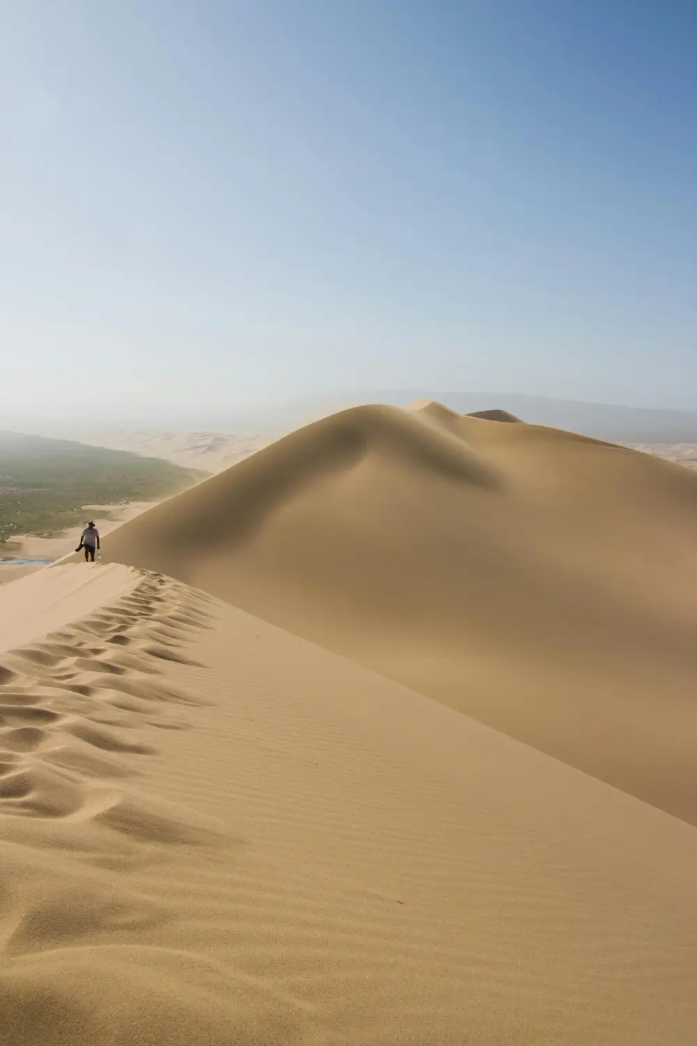 A lone hiker walking along the sharp ridgeline of a towering golden sand dune with misty desert valleys below