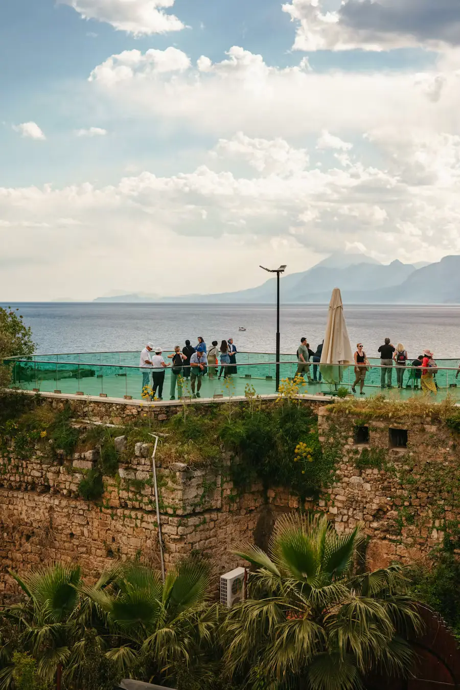 People standing atop an old stone fortress wall overlooking the sea, with mountains visible across the water under a partly cloudy sky.
