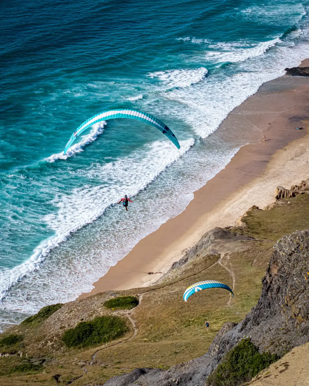 Paragliders soar above turquoise waves and a sandy beach bordered by rocky coastal cliffs.