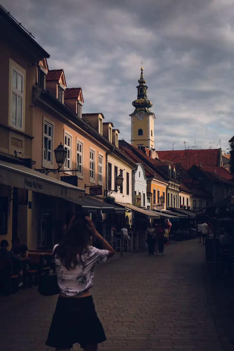 Person walking down a cobblestone European street lined with pastel-colored buildings, a clock tower visible in the distance under a cloudy sky.