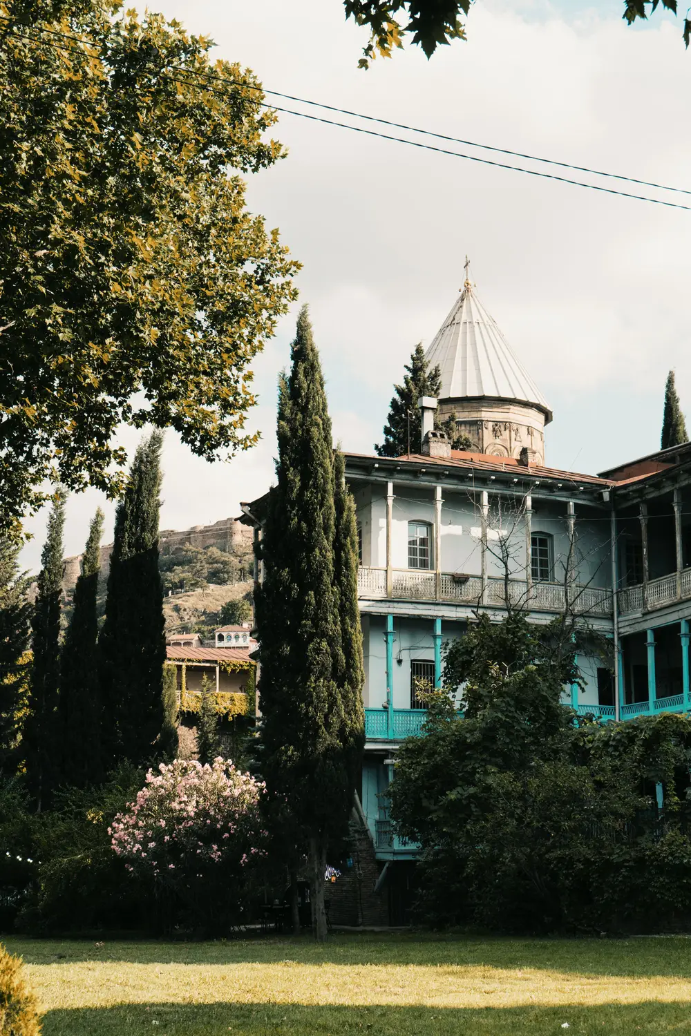 A historic turquoise-and-white building with a conical roof, surrounded by tall cypress trees and a lush garden.