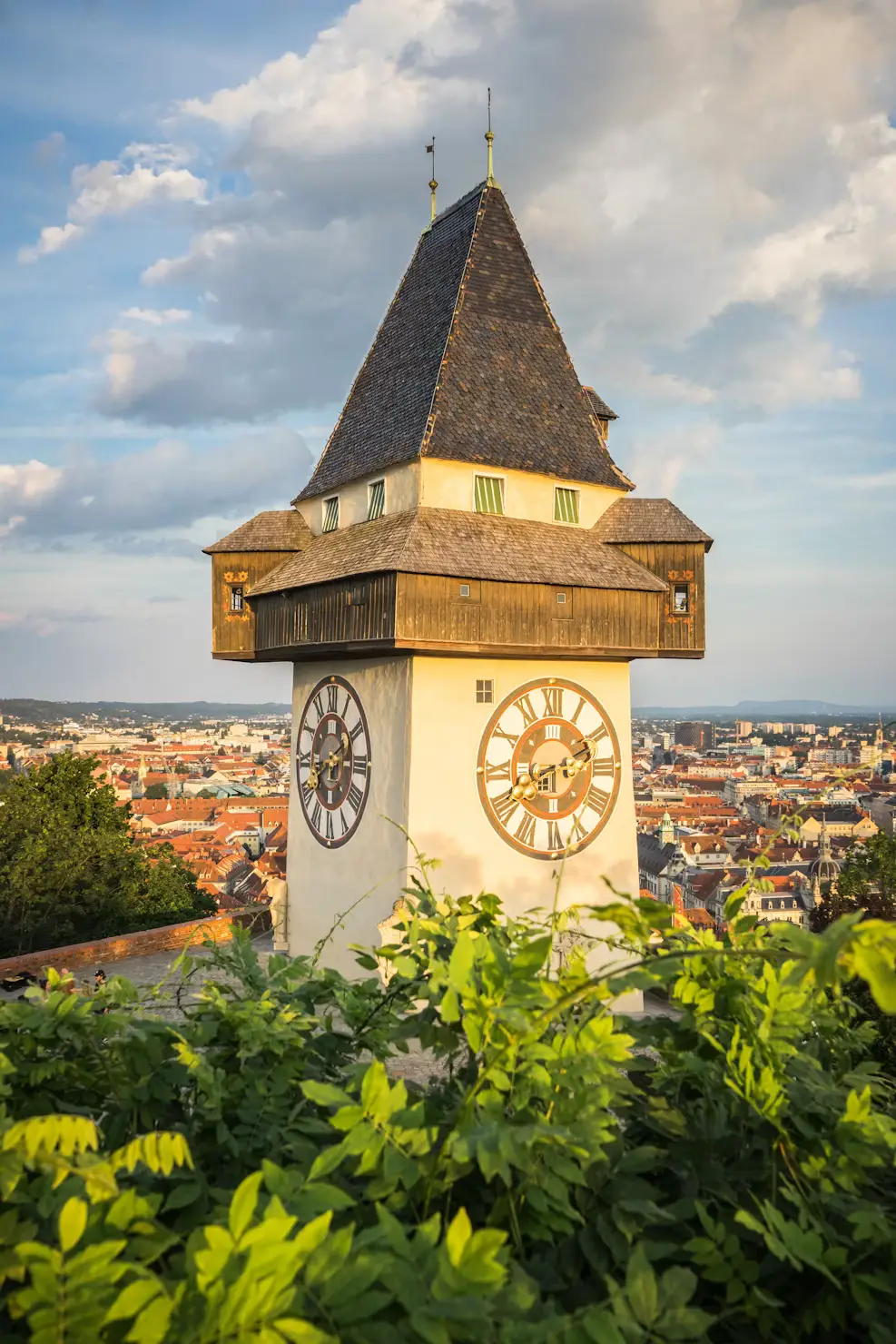 A clock tower with a steep pyramidal roof rises above greenery, overlooking a city skyline at sunset.