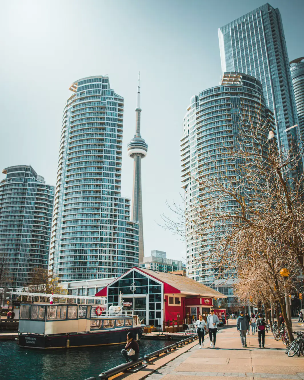 Toronto's CN Tower and skyscrapers rising above the waterfront, with people walking along the harbor promenade.