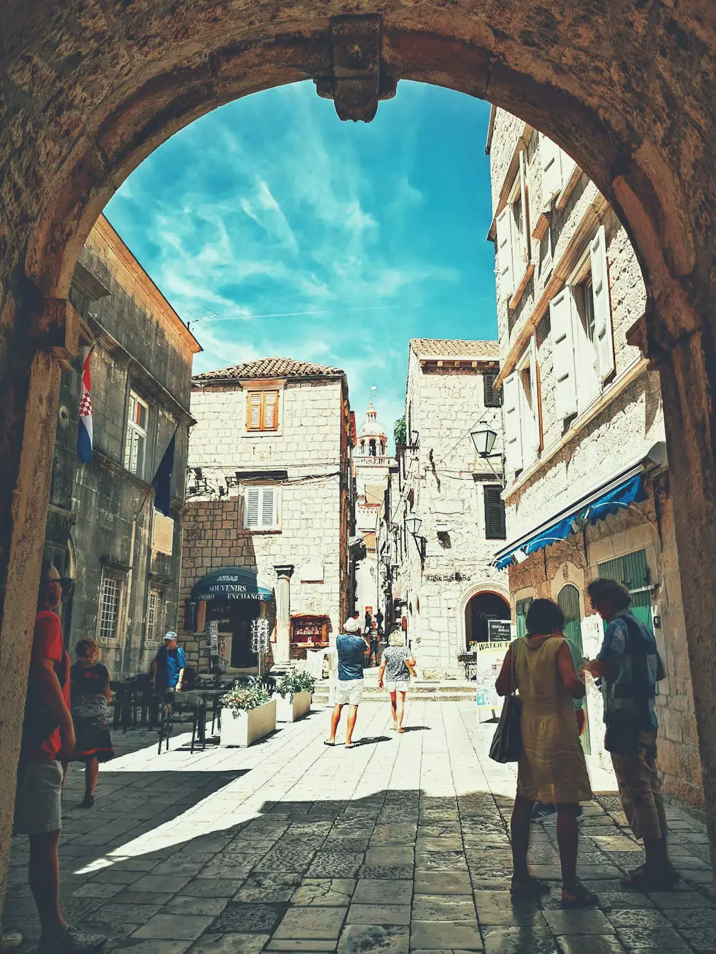 View through a stone archway into a sunlit cobblestone street with white buildings and pedestrians.