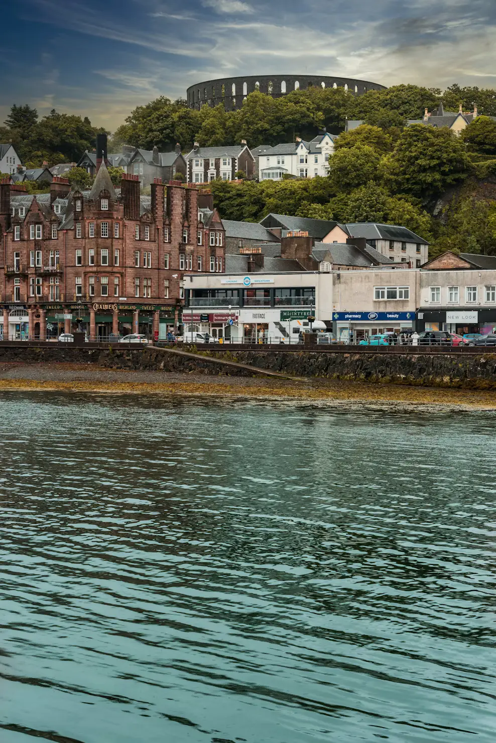 Waterfront buildings of Oban along the shore, with McCaig's Tower crowning the green hillside above the bay.