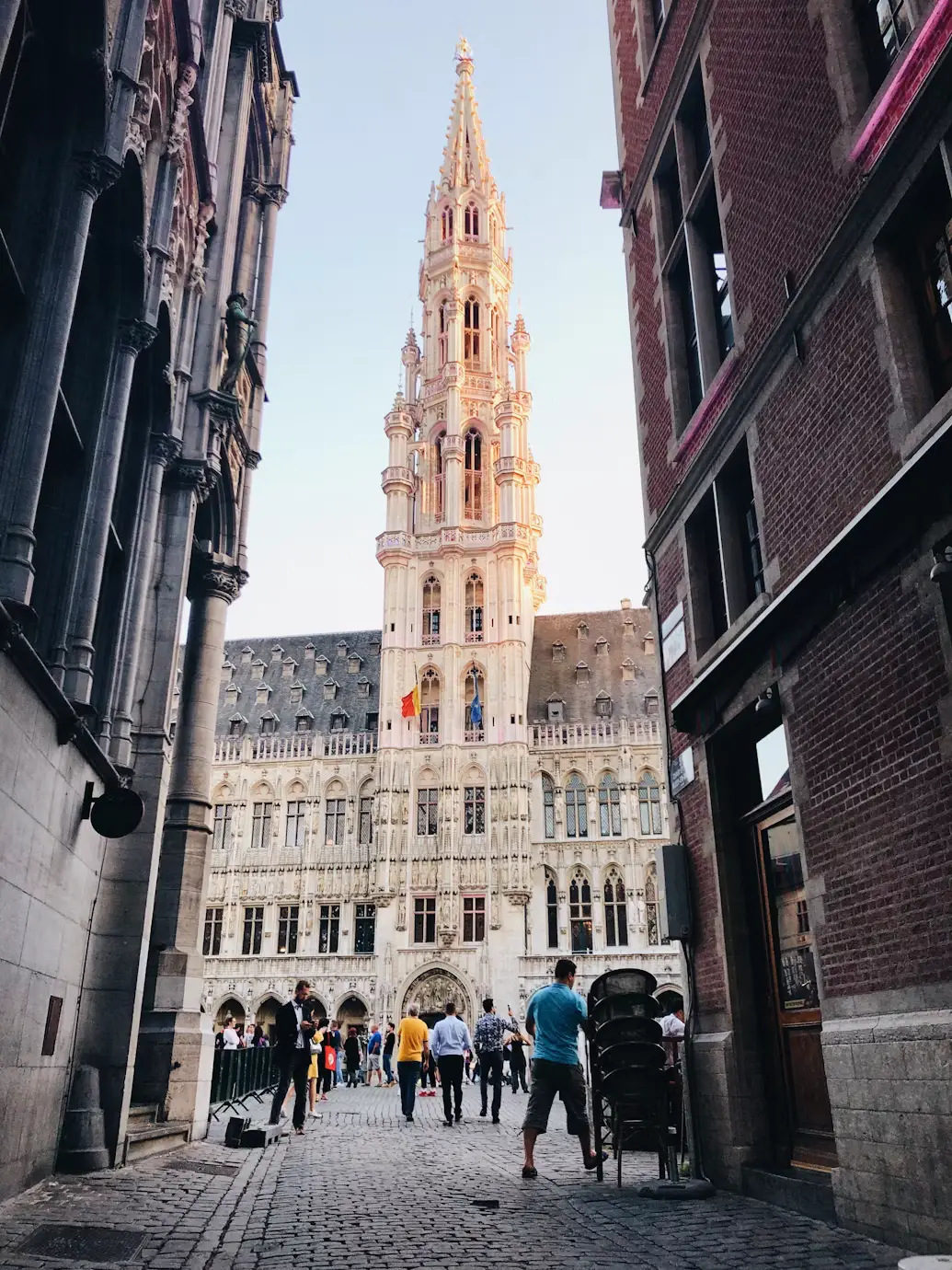 View down a narrow alley toward the illuminated spire of Brussels' Town Hall at dusk, with people walking below.