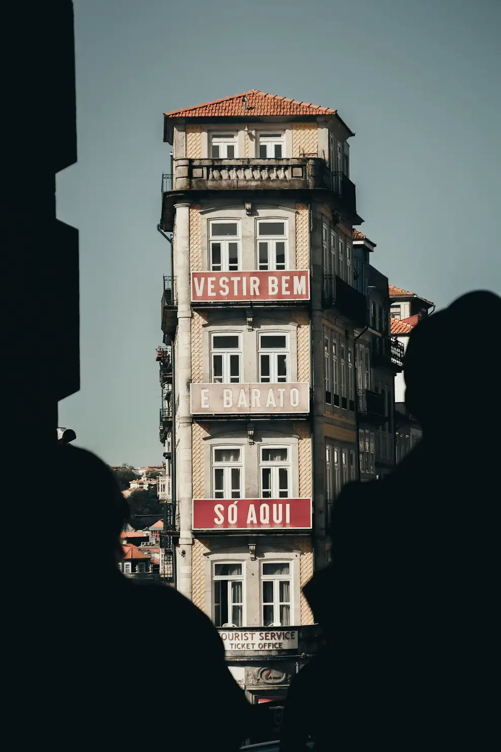 A narrow multi-story building with red signs reading “Vestir Bem e Barato Só Aqui,” framed by silhouetted pedestrians.