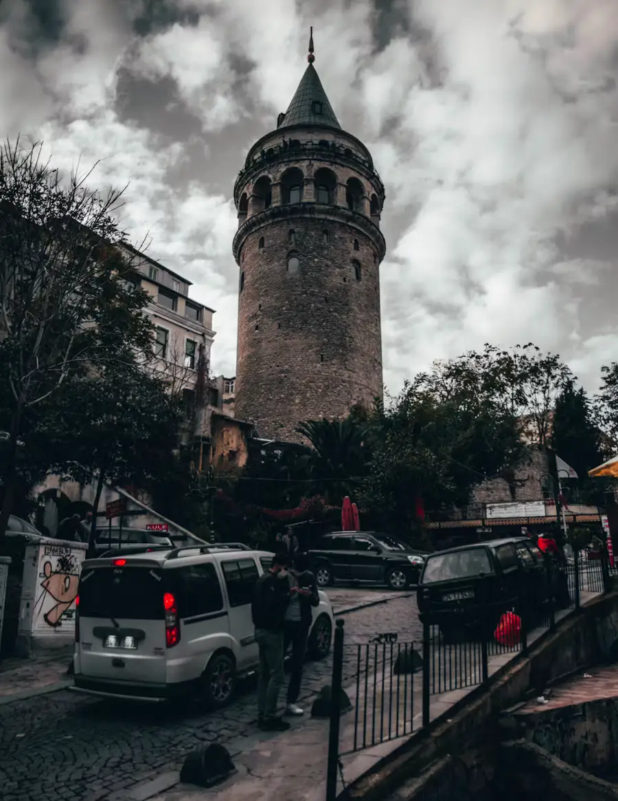 The stone Galata Tower rising above a cobblestone street with parked cars, under a dramatic cloudy sky.