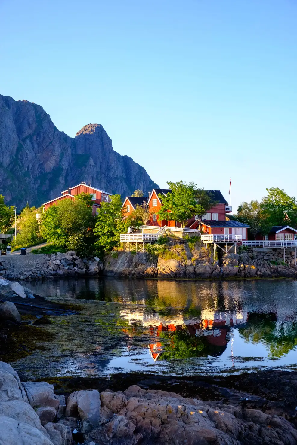 Red waterfront cabins reflected in a calm rocky cove, with a steep mountain peak rising behind under a clear evening sky.
