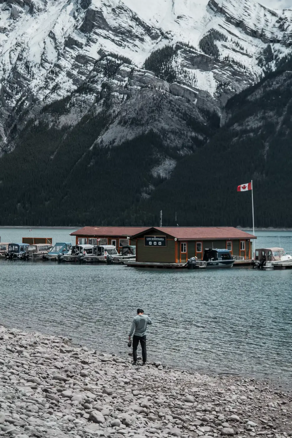 A person standing on a rocky lakeshore facing snow-capped mountains, with small cabins visible along the water's edge.